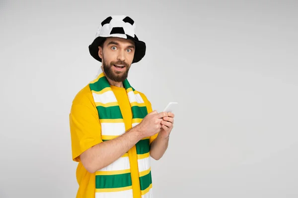 Hombre positivo y curioso en el sombrero del ventilador de fútbol y bufanda a rayas que sostiene el teléfono inteligente y mirando a la cámara aislada en gris - foto de stock