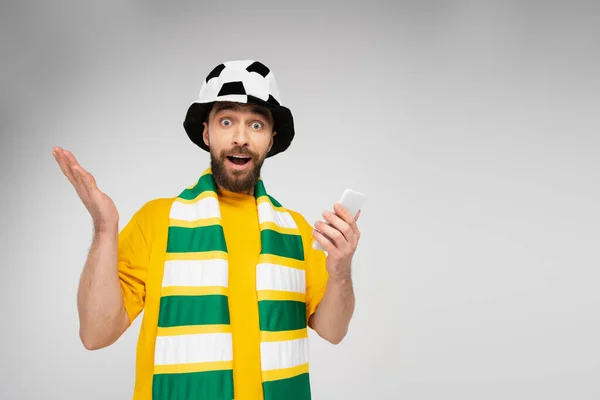 Удивленный человек в полосатом шарфе и футбольной шляпе болельщика держа сотовый телефон и глядя на камеру изолированы на серый — стоковое фото