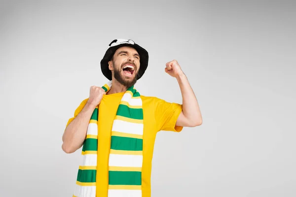 Homem feliz com cachecol listrado e chapéu de fã de futebol gritando e mostrando gesto triunfo isolado no cinza — Fotografia de Stock