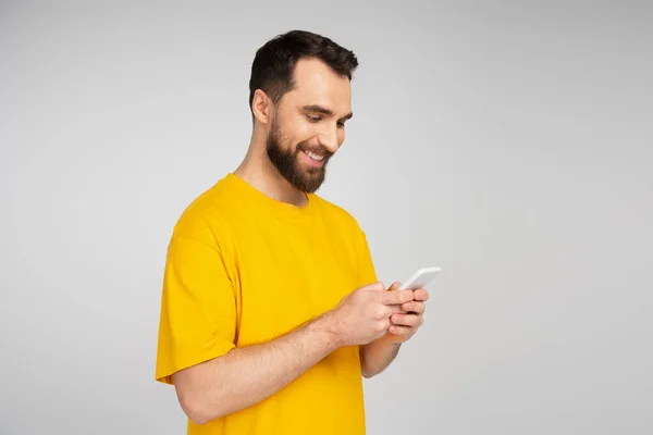 Hombre barbudo sonriente en camiseta amarilla usando teléfono móvil aislado en gris - foto de stock