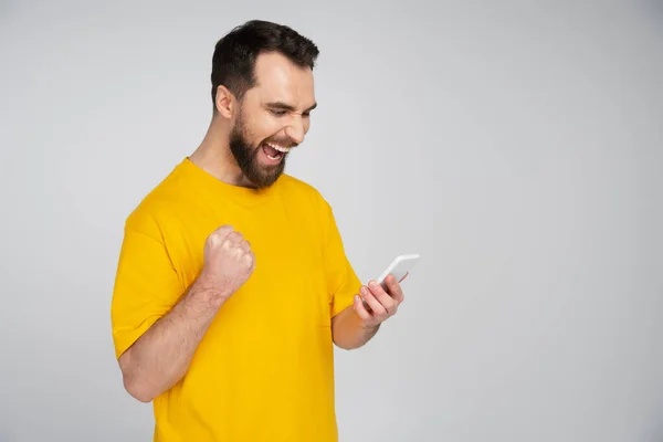 Homem barbudo animado com smartphone mostrando gesto de vitória e gritando isolado no cinza — Fotografia de Stock