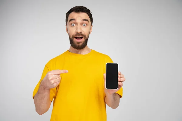 Amazed bearded man in yellow t-shirt pointing at smartphone with blank screen isolated on grey — Stock Photo