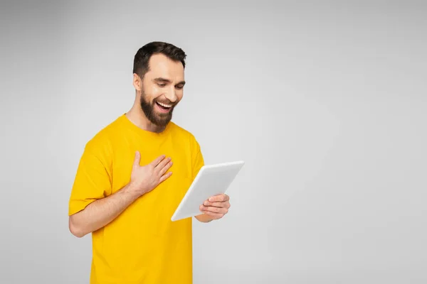 Смеющийся мужчина в желтой футболке трогает грудь, глядя на цифровой планшет, изолированный на сером — стоковое фото