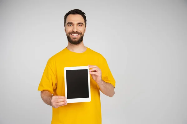 Hombre alegre con barba mostrando tableta digital con pantalla en blanco mientras sonríe a la cámara aislada en gris - foto de stock
