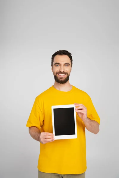 Брюнетка бородатий чоловік показує цифровий планшет з порожнім екраном і посміхається на камеру ізольовано на сірому — стокове фото