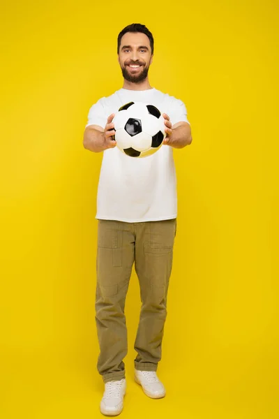 Full length of happy bearded man in white t-shirt and beige pants holding soccer ball in outstretched hands on yellow background — Stock Photo