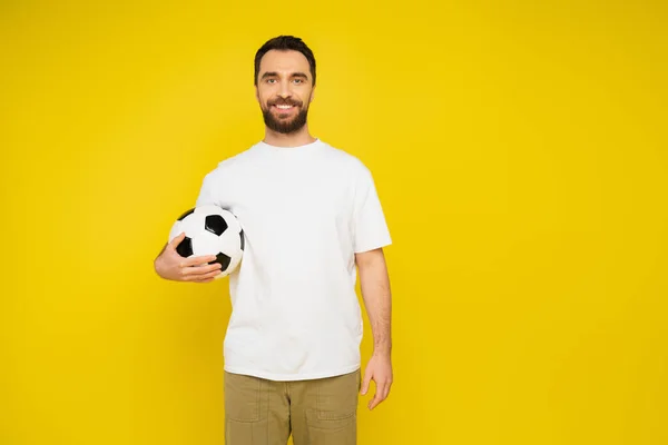 Hombre barbudo feliz en camiseta blanca de pie con pelota de fútbol y mirando a la cámara aislada en amarillo - foto de stock