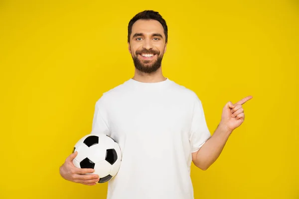 Abanico deportivo barbudo en camiseta blanca sosteniendo pelota de fútbol y señalando con el dedo aislado en amarillo - foto de stock