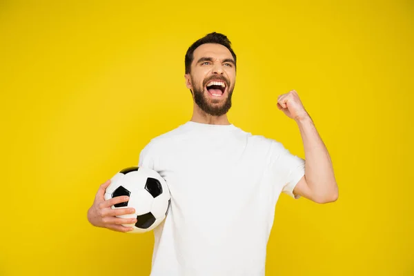 Fanático del fútbol lleno de alegría con pelota de fútbol gritando y mostrando gesto de victoria aislado en amarillo - foto de stock