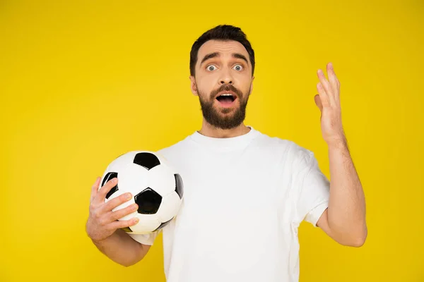 Escandalizado y frustrado fanático de los deportes sosteniendo pelota de fútbol y gestos aislados en amarillo — Stock Photo