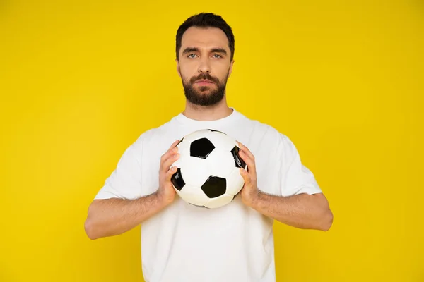 Серйозний бородатий спортивний вентилятор у білій футболці тримає футбольний м'яч і дивиться на камеру ізольовано на жовтому — стокове фото