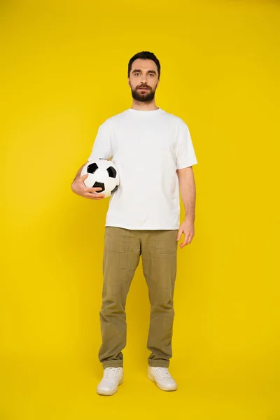 Piena lunghezza di bruna uomo barbuto in bianco t-shirt e pantaloni beige in piedi con pallone da calcio su sfondo giallo — Foto stock