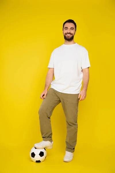 Full length of cheerful man in beige pants and white t-shirt stepping on soccer ball on yellow background — Stock Photo