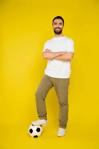 Volle Länge des lächelnden bärtigen Mannes, der mit verschränkten Armen auf gelbem Hintergrund auf den Fußball tritt — Stockfoto