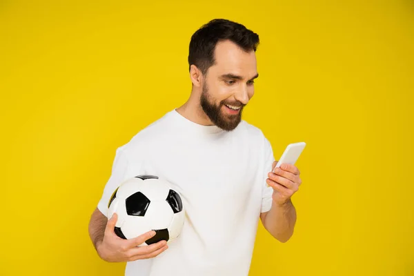 Fan de football souriant tenant ballon de football tout en regardant le jeu sur smartphone isolé sur jaune — Photo de stock
