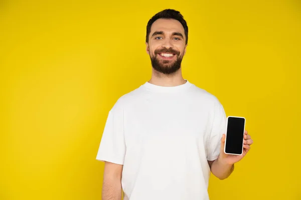 Весёлый бородатый мужчина в белой футболке, держащий смартфон и смотрящий на камеру, изолированную на жёлтом — стоковое фото