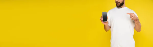 Vista ritagliata dell'uomo barbuto in t-shirt bianca che punta al cellulare con schermo bianco isolato su giallo, banner — Foto stock