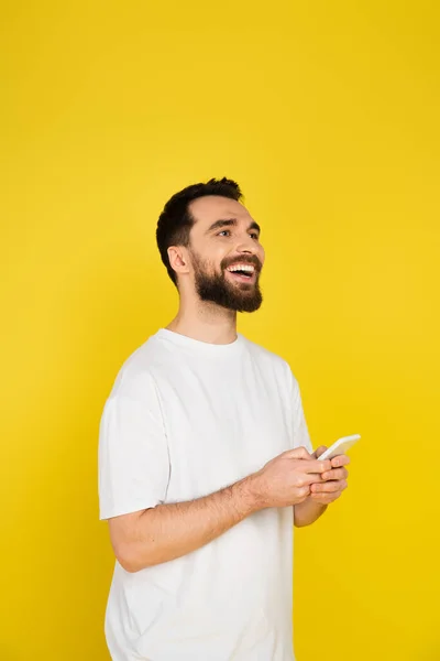 Hombre barbudo excitado en camiseta blanca sosteniendo el teléfono móvil mientras mira hacia otro lado aislado en amarillo - foto de stock