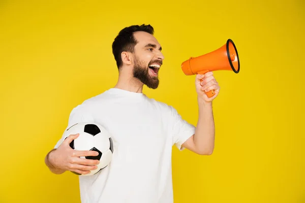 Abanico de deportes barbudos en camiseta blanca sosteniendo pelota de fútbol y gritando en megáfono aislado en amarillo - foto de stock