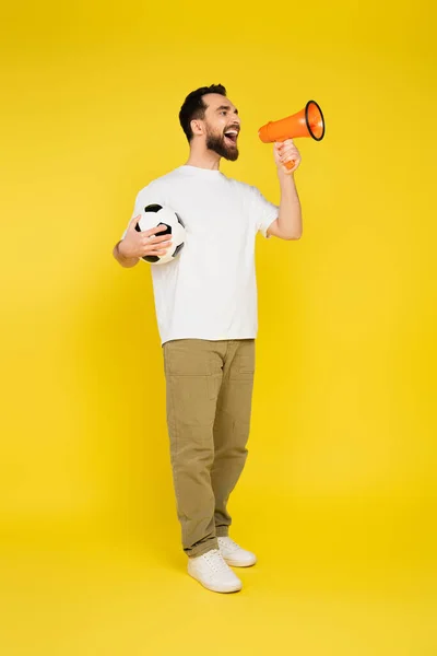 Full length of man in white t-shirt and beige pants holding soccer ball and screaming in megaphone on yellow background — Stock Photo