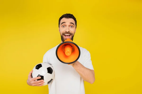 Admirador de los deportes sorprendidos con pelota de fútbol gritando en megáfono y mirando a la cámara aislada en amarillo - foto de stock