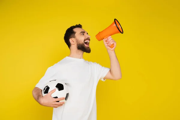 Ventilador de futebol animado gritando em alto-falante enquanto segurando bola isolada no amarelo — Fotografia de Stock