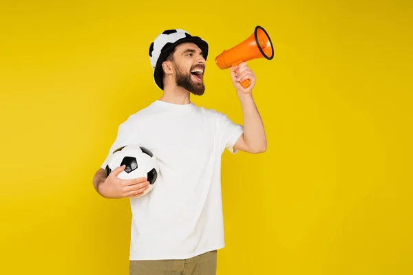 Взволнованный спортивный фанат в шляпе, стоящий с футбольным мячом во время крика в громкоговорителе изолированы на желтый — стоковое фото