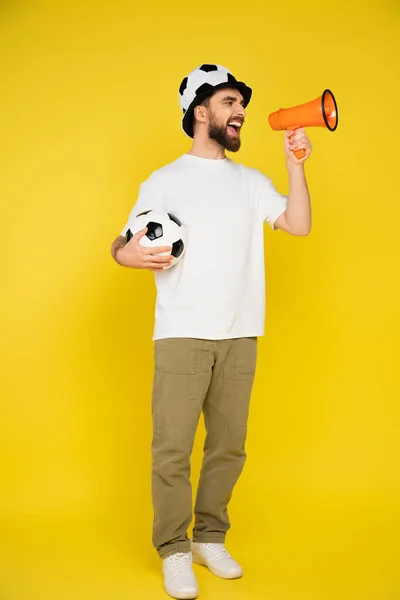 Sportfan in voller Länge mit Hut hält Fußball in der Hand und schreit im Megafon auf gelbem Hintergrund — Stockfoto