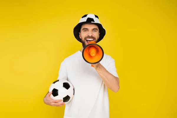 Thrilled man in football fan hat standing with soccer ball and shouting in loudspeaker isolated on yellow — Stock Photo