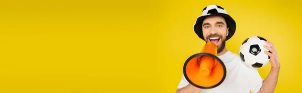Uomo gioioso in cappello tifoso di calcio che tiene palla da calcio e urla in megafono isolato sul giallo, striscione — Foto stock