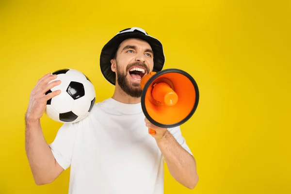 Vista en ángulo bajo del ventilador deportivo con pelota de fútbol gritando en megáfono aislado en amarillo - foto de stock