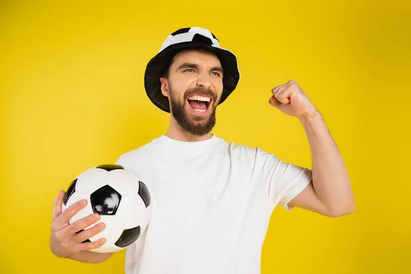 Кричащий футбольный фанат в шляпе, держащий футбольный мяч и показывающий успешный жест изолирован на желтый — стоковое фото
