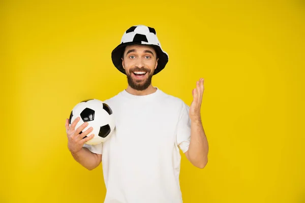 Hombre barbudo feliz en el ventilador de fútbol sombrero sosteniendo pelota de fútbol y mirando a la cámara aislada en amarillo - foto de stock
