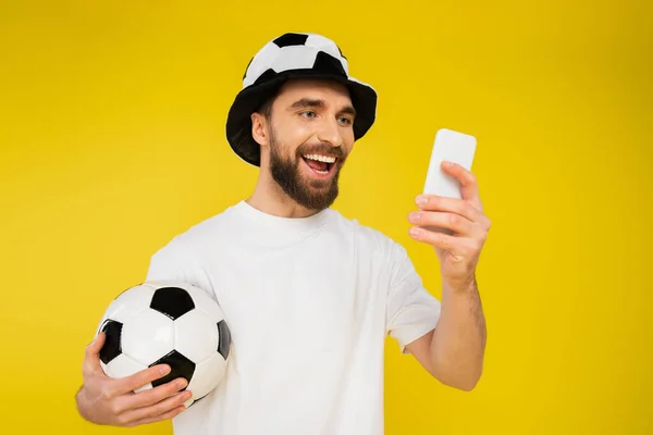Tifoso sportivo soddisfatto in cappello guardando partita di calcio su smartphone isolato su giallo — Foto stock