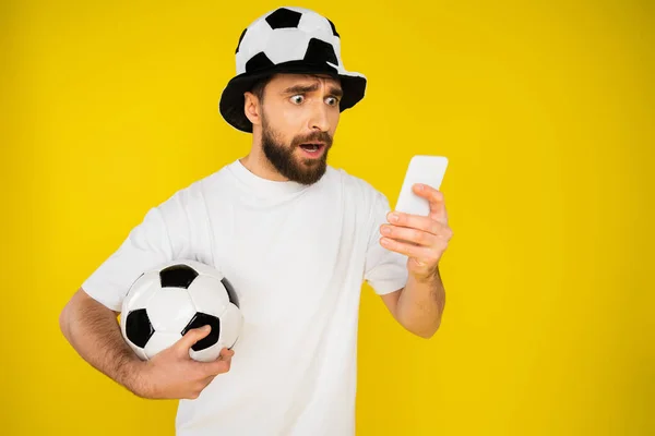 Хвилюючий фанат футболу в капелюсі дивиться гру на мобільному телефоні ізольовано на жовтому — стокове фото