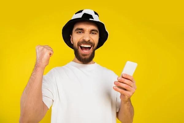 Alegre ventilador de fútbol en sombrero mostrando gesto de triunfo y la celebración de teléfono celular aislado en amarillo - foto de stock