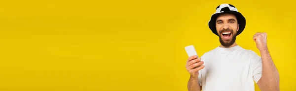 Crier homme en chapeau de ventilateur de football tenant smartphone et montrant geste de victoire isolé sur jaune, bannière — Photo de stock