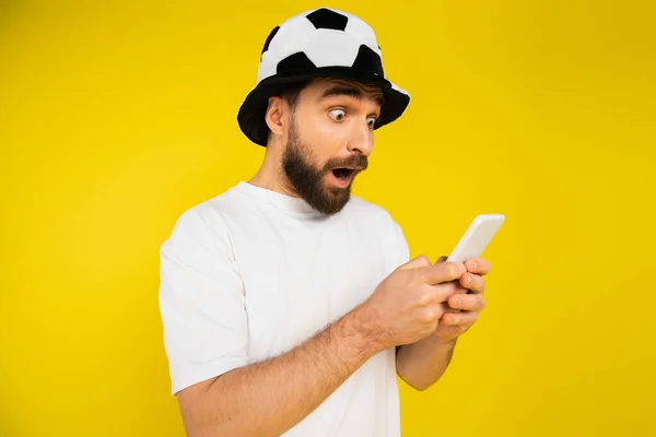 Sorprendido hombre en fútbol ventilador sombrero viendo campeonato en el teléfono celular aislado en amarillo - foto de stock