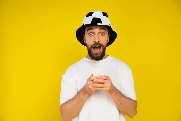 Homme étonné en chapeau de ventilateur de football tenant le téléphone portable et regardant la caméra isolée sur jaune — Photo de stock