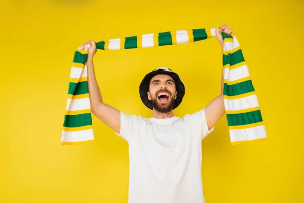 Joyful man in football fan hat shouting while holding striped scarf in raised hands isolated on yellow — Stock Photo