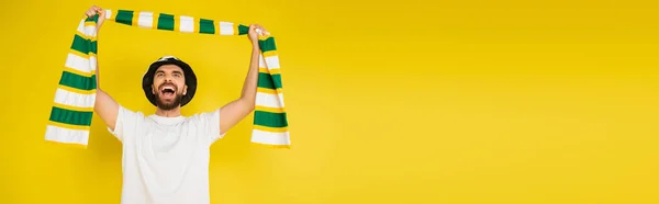 Emocionado ventilador deportivo sosteniendo bufanda a rayas en las manos levantadas y gritando aislado en amarillo, pancarta - foto de stock