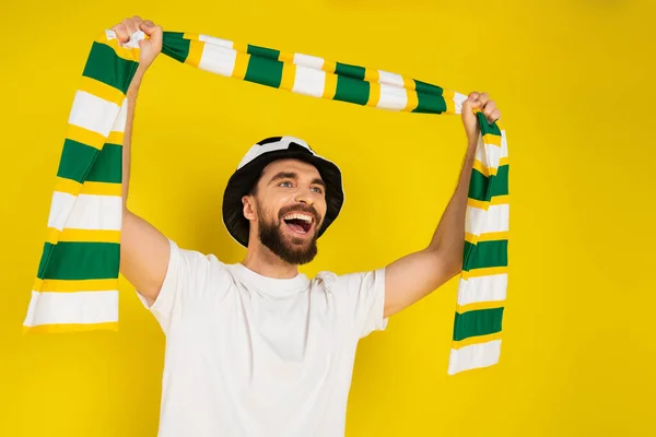 Восторженный спортивный болельщик в шляпе кричит, держа полосатый шарф в поднятых руках изолированы на желтый — стоковое фото