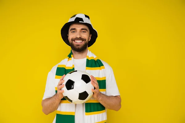 Счастливый человек в шляпе футбольного болельщика и полосатый шарф держа футбольный мяч изолирован на желтый — стоковое фото