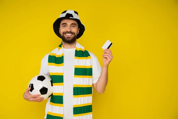 Alegre fanático del fútbol con balón de fútbol y tarjeta de crédito mirando a la cámara aislada en amarillo - foto de stock