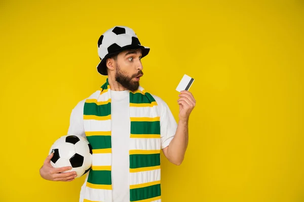 Erstaunter Mann mit Fußballfan-Mütze und gestreiftem Schal, der Fußballball in der Hand hält und die Kreditkarte isoliert auf gelb betrachtet — Stockfoto