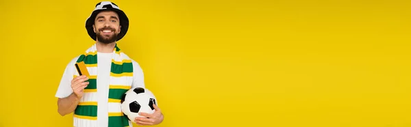 Счастливый человек в шляпе футбольного болельщика стоя с кредитной картой и футбольный мяч изолирован на желтый, баннер — стоковое фото