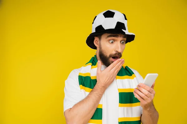 Schockierter Mann mit Fußballfan-Mütze verdeckt offenen Mund mit der Hand, während er das Smartphone isoliert auf gelb betrachtet — Stockfoto