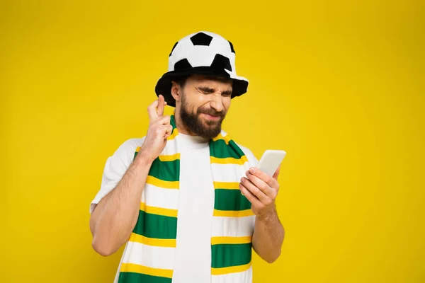 Напружений футбольний вентилятор із закритими очима, тримаючи схрещені пальці та мобільний телефон ізольовані на жовтому — стокове фото