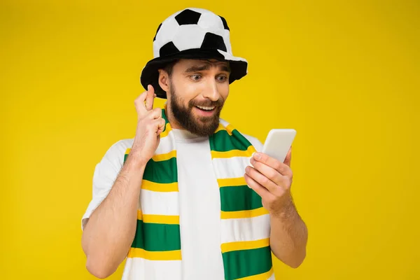 Homem tenso em chapéu de fã de futebol olhando para smartphone e segurando dedos cruzados isolados no amarelo — Fotografia de Stock