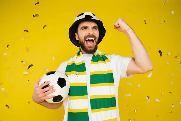 Захоплений спортивний вентилятор з футбольним м'ячем, що кричить і показує жест виграшу під падінням конфетті на жовтому тлі — стокове фото